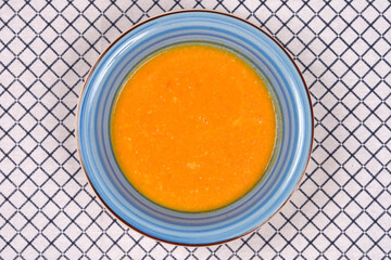 An overhead shot of creamy orange carrot and peanut soup in a blue ceramic bowl placed on a white...