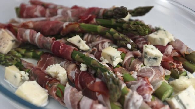 Bacon-coated asparagus spears with gorgonzola cubes placed in a clear oven dish on white.