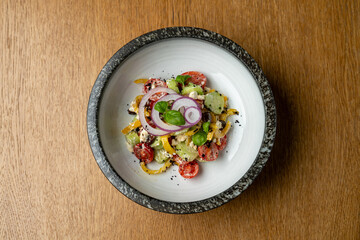 A vibrant salad featuring cherry tomatoes, onions, cucumbers, and herbs arranged in a stylish bowl rests on a wooden table. Ideal for enjoying al fresco dining