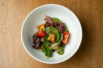A colorful garden salad is served in a white bowl, showcasing a mix of vibrant greens, ripe tomatoes, and roasted vegetables. The dish glistens with a light dressing