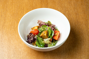 A vibrant garden salad features fresh greens, ripe tomatoes, and crispy toppings. The dish is beautifully arranged in a sleek white bowl against a warm wooden background