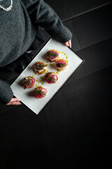 A person in a cozy sweater presents a platter of beautifully crafted canapes adorned with colorful garnishes. The dimly lit atmosphere highlights the elegance of the dish, inviting guests to indulge