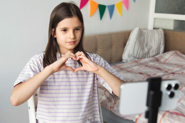 girl recording a video at home, forming a heart gesture with her hands. Positive child vlogging, expressing love and support while creating digital content in a cozy, bright bedroom setting.