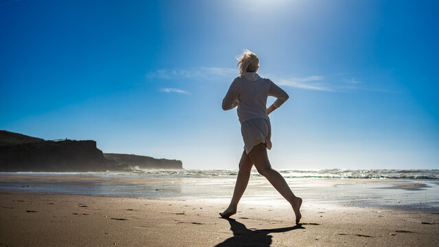 Beautiful middle-aged woman running along ocean on sandy Bordeira beach in Portugal on sunny summer day. Side view	