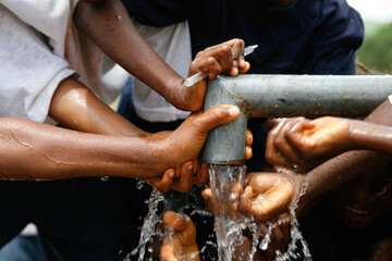 School with a biro pen and children's hands and water drops splashing beneath a running water tap and metal pipe in a West African village as they drink the clean water from a bore hole