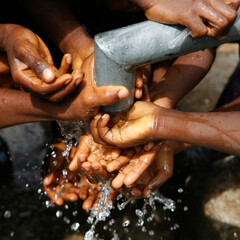 Children's hands and water drops splashing beneath a running water tap in a West African village as...