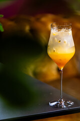 A vibrant, tropical mango passion fruit cocktail in an elegant stemmed glass with ice and seeds on a dark table, featuring a warm bokeh background