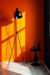 Modern black tripod floor lamp casting dramatic shadows against a vibrant, warm orange wall in a minimalist interior space