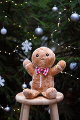 A cute gingerbread plush toy standing in front of a beautifully decorated Christmas tree, surrounded by warm holiday lights and festive ornaments. The cozy Christmas atmosphere highlights the playful.