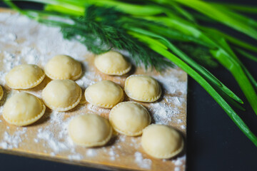 dumplings, not boiled, freshly molded on a kitchen board sprinkled with flour. Greens of spices along the edge of the frame. on a black background. Shooting angle from top to side. High quality photo