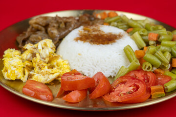 A balanced meal on a brass plate featuring white rice topped with sauce, stir fried beef slices, scrambled eggs, sliced tomatoes, and sautEed green beans and carrots against a red background.