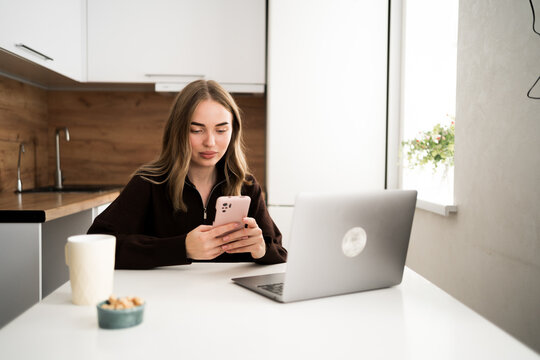 Young woman checking laptop typing message on phone in a kitchen at home - Powered by Adobe