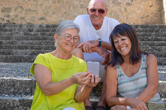 Three smiling seniors sit together on outdoor stone steps, happily looking at a smartphone. The scene conveys friendship, connection, leisure, and the joy of sharing moments through technology