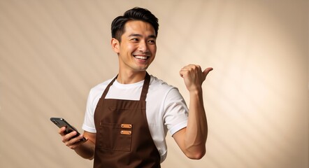 Cheerful asian barista or shop owner pointing at copy space. Happy male entrepreneur in an apron holding a smartphone and smiling. Small business and customer service concept