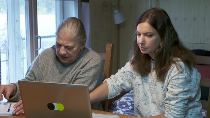 A focused daughter helps her concerned mother navigate a laptop screen while sitting at a wooden table. They analyze digital information together in a rustic home setting. - Powered by Adobe