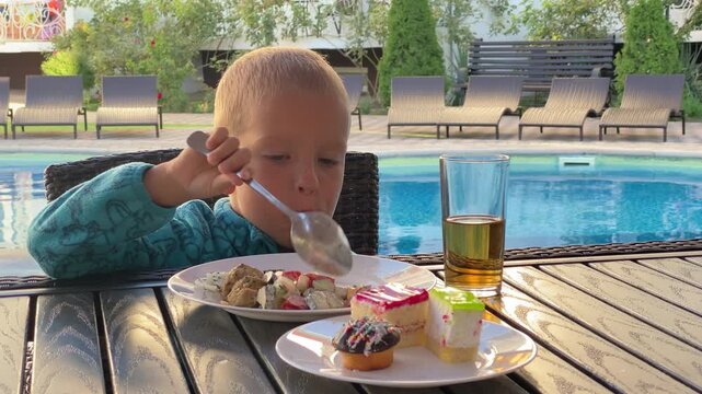 A hungry boy eats dinner with a spoon at a poolside table. The kid enjoys a meal and sweet desserts near the blue water during a sunny vacation day.
