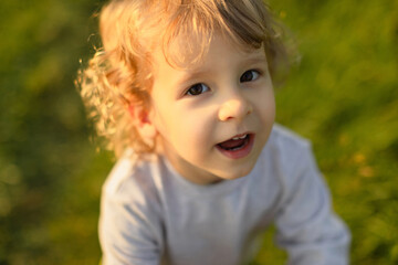 Close-up portrait of a smiling toddler in warm evening light, looking up with bright eyes and natural expression. Soft green background and authentic childhood moment.