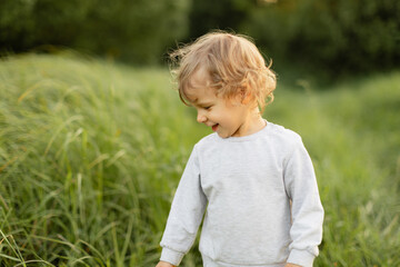 Smiling toddler walking through tall grass on a sunny day, looking down with a joyful natural expression. Soft light and candid outdoor childhood moment.