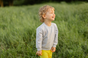Smiling toddler standing in tall green grass on a sunny day. Candid outdoor moment with warm light, joyful expression and natural childhood energy.