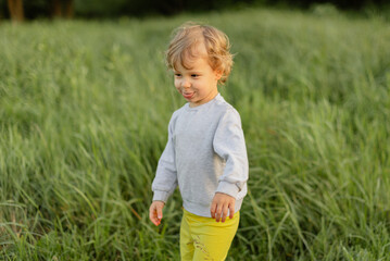 Playful toddler walking through tall grass, making a funny face in soft natural light. Candid outdoor moment showing real childhood personality and movement.
