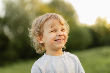 Smiling blonde toddler enjoying a sunny outdoor moment, captured in soft natural light with a blurred green background. Warm, joyful childhood expression.