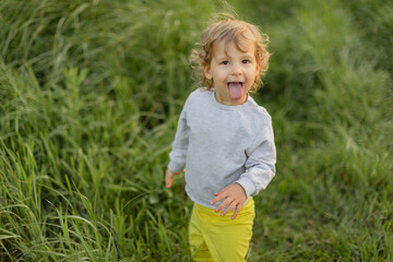 Playful toddler sticking out tongue while walking through tall green grass. Candid outdoor moment full of energy, natural light and real childhood expression.