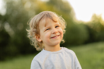 Smiling blonde toddler outdoors in soft natural light, captured with a warm candid expression against a blurred green background. Happy childhood moment.