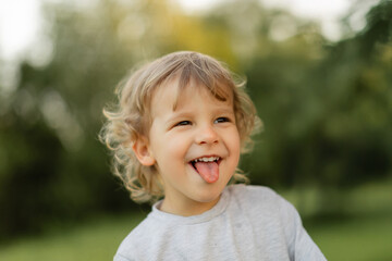 Playful blonde toddler sticking out tongue while smiling outdoors, captured in soft natural light with blurred green background. Joyful childhood moment and authentic candid expression.