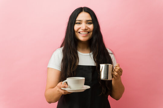 Smiling young female barista holding cup and frothed milk pitcher