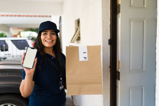 Smiling delivery woman holding a package and smartphone with blank screen at a residential front door