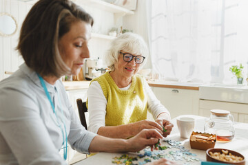 Caregiver in geriatric clinic or retirement home helping senior lady with puzzle as treatment for dementia rehabilitation, both sitting in kitchen at desk and playing board games together
