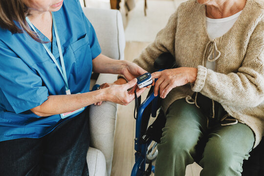 Image of two unrecognizable people, nurse in blue uniform doing saturation assessment and checking pulse with finger oximeter to check oxygen level of elderly woman in wheelchair at geriatric clinic