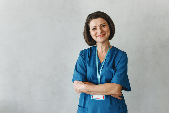 Successful confident Caucasian female massage therapist or kinesiotherapist posing against grey studio background with copy space for your advertising content in blue uniform standing with folded arms