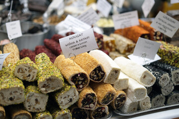 Multiple nougat rolls lie stacked on metal trays. Their surfaces show pistachios, nuts, shredded pastry and powdered coatings. Small labels with text stand among the rows.