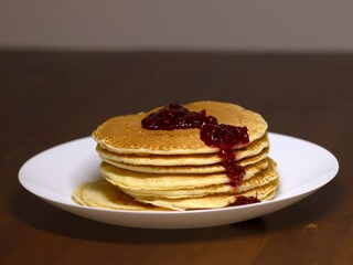 A pile of fresh pancakes with raspberry jam on top, on a white plate. Standing on a wooden brown table.