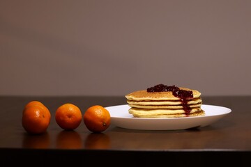 Many pancakes on a white plate, raspberry jam on top, a few tangerines on the side. All this on an old brown table