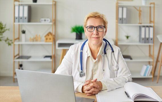 Mature female doctor smiling at desk, medical consultant, female medical nurse or practitioner, clinic manager analyzing data, keeping patient information, health center assistant, client advice