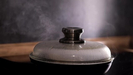 Frying large meat sausages in a frying pan under a lid, steam comes out from under the lid, the cook lifts the lid.