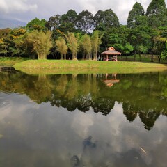 Fototapeta premium A traditional gazebo on a grassy bank is perfectly reflected in a calm, dark lake. Surrounded by lush bamboo and evergreen trees under a cloudy sky.