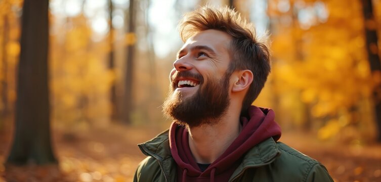 Young man with beard smiles looking up in golden autumn forest. Person enjoys nature, fall season walk, outdoor recreation and good weather. Male person happy, casual style, relaxed.