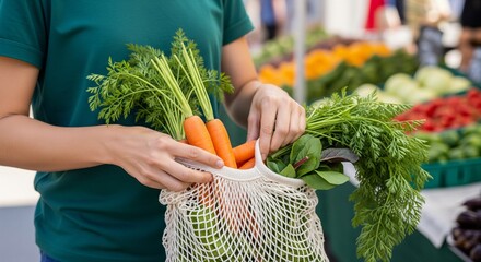 A person in a green shirt puts a bunch of fresh carrots into a reusable mesh grocery bag at an outdoor farmers' market.