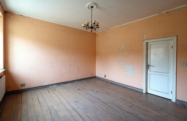 Fototapeta premium Empty old room with peeling peach paint walls, dark wooden floor. Vintage chandelier from ceiling. White closed door, radiator visible. Room dirty, abandoned, shows decay, long term neglect. Poor