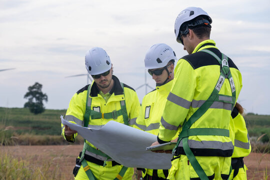 Workers discuss plans while inspecting wind turbine site in bright yellow safety gear during daylight hours - Powered by Adobe