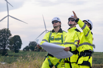 Workers review plans at a wind farm site as turbines spin in the background during a clear day
