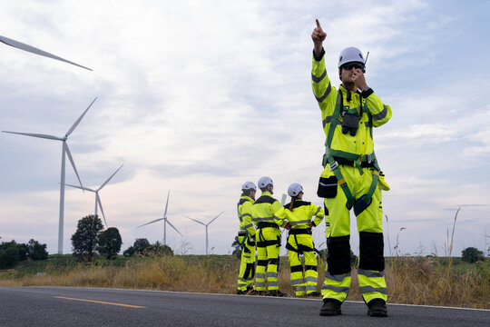 Workers in bright uniforms overseeing wind turbine maintenance in a rural area during cloudy afternoon