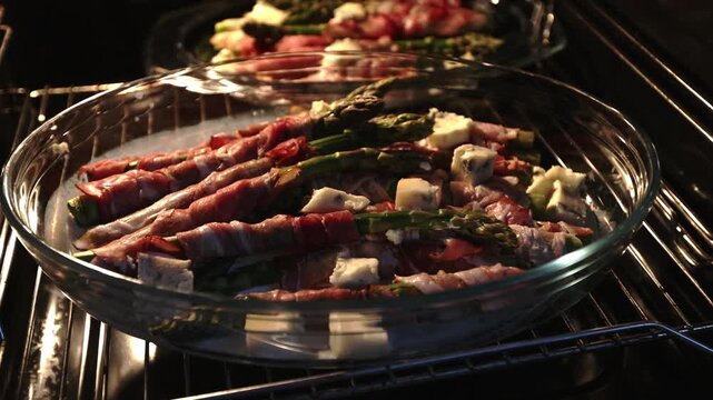 Bacon-wrapped green asparagus with gorgonzola cubes in a clear baking dish filmed inside a heated oven.