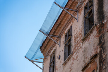 Old damaged building facade with broken windows and protective metal mesh installed above to prevent falling debris and ensure safety during planned renovation.