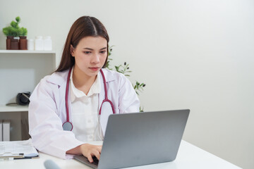 Asian female doctor working on laptop at clinic office, reviewing patient files and entering data into system, professional medical staff using digital tools for online healthcare and diagnosis