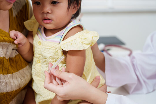 Pediatric nurse checks toddler temperature with thermometer before injection, asian mother comforts baby during clinic visit, healthcare routine shows gentle professional care for child wellness