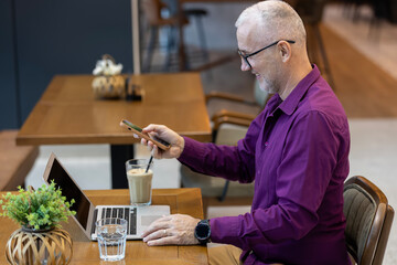 Mature man smiling using smartphone with laptop at cafe